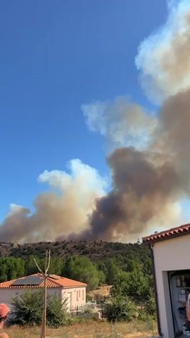 A wildfire is visible on a hillside in the distance, emitting a large plume of smoke that rises into the clear blue sky. In the foreground, there are two houses with orange tiled roofs. To the left, a white and pink house with solar panels on its roof. To the right, a white structure, possibly a garage, with an electrical panel visible. Three people are standing in the foreground, looking towards the fire. One person on the left, wearing a red cap, a dark tank top, and shorts. In the center, a person from the back, wearing a blue shirt. To the right, another person from the back, wearing a blue shirt. The ground in the foreground is covered with gravel. The overall scene is one of a community observing a natural disaster.