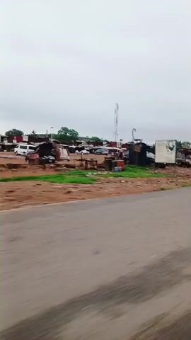 The video captures a roadside scene in Karshi, Nigeria, filmed from a moving vehicle, likely an auto-rickshaw given the visible right-side mirror at the 0:02 mark. The time of day is daytime, under an overcast sky, suggesting cloudy weather.

The scene features a paved road with some visible wear, along which the vehicle is traveling. To the right of the road, there's an unpaved area of reddish-brown earth mixed with patches of green vegetation. This area is populated with numerous informal structures, resembling shacks or small businesses, constructed from materials such as corrugated metal and wood. Several cars, varying in model and condition, are parked irregularly among these structures; some appear functional while others might be part of a mechanics' area or used car lot. A few green trees are scattered throughout this roadside development.

As the vehicle moves, a black car passes by in the opposite direction at approximately 0:03. The overall impression is that of a bustling, semi-urban or rural-urban fringe environment. No clear, readable text is visible on any signs or objects throughout the video.