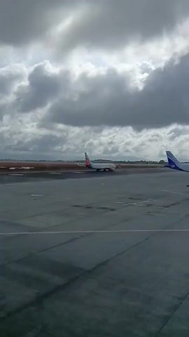 The video shows a gray tarmac on a cloudy day, with two airplanes visible. In the distance, an airplane with a white body and red accents is moving along the runway. Closer to the camera is another airplane, with a white body and blue tail, sitting stationary on the tarmac.