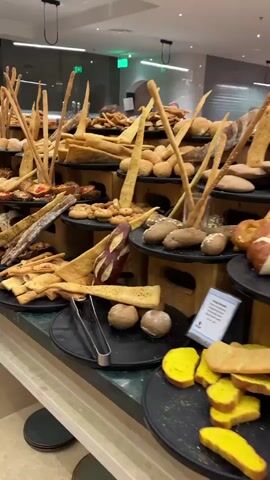 The camera slowly pans across a multi-tiered display of breads and pastries. The various types of breads are arranged on round, black serving plates atop wooden risers.  There are long breadsticks, thin triangular flatbreads, small rolls, sesame breads, pizzas, and open-faced sandwiches.  A pair of tongs rest on one of the plates. The display is well-lit and is likely at a buffet.
