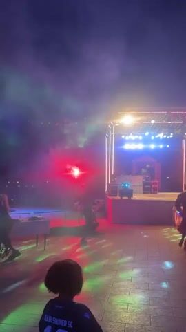 A nighttime outdoor event features a raised stage with a DJ setup and colorful lights. Several individuals are dancing in front of the stage, illuminated by stage lights and smoke. One person in the foreground, facing away from the camera, is watching the event. Red and white lights are visible on the left side of the scene.