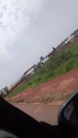 The video shows a view from inside a vehicle, looking out the passenger side window. The camera is angled, making the scene appear tilted. The surroundings are rural, with a dirt road in the foreground and a gas station with a red canopy visible in the middle ground. Beyond the gas station, there are buildings with pitched roofs and trees. The sky is overcast. The vehicle appears to be moving, as suggested by the slight blur and the changing perspective. There are no visible people in the frame.