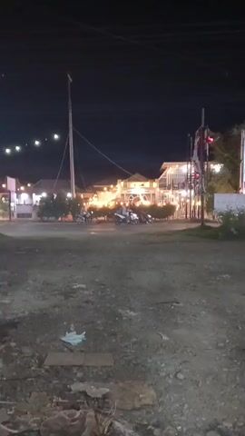 A nighttime view of a dirt lot in the foreground with trash scattered on the ground. In the background, a street with a few motorcycles is visible, along with buildings that appear to be restaurants or shops with lights on. A tall pole with wires extends upwards on the left. A blue truck drives across the street from left to right, its lights illuminated. A traffic light with a red signal is visible on the right side of the frame.