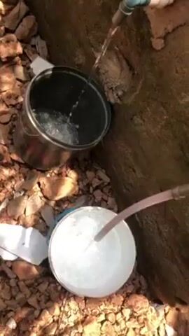 The video shows water being poured into two separate containers. A metal bucket is being filled with water coming directly from a pipe, while a white bucket is being filled by water flowing through a pink tube attached to a faucet. The containers are placed on a rocky, outdoor surface. The background consists of a textured, earthen wall.