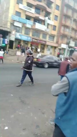 The video shows a bustling street scene in what appears to be a busy urban area. The camera is positioned from the perspective of someone standing on the side of the road, capturing the lively activity of pedestrians and vehicles.

In the foreground, several individuals can be seen walking across the street. These include men and women, dressed in casual clothing. One man is wearing a blue vest, his arms crossed, seemingly observing the scene. Some of the pedestrians are wearing masks, possibly in response to health concerns.

The street itself is a moderately wide paved surface with some signs of wear and tear. Along the sidewalks lining the street are various commercial establishments, with signs indicating shops, supermarkets, and offices. The buildings are generally multi-story, with a mix of architectural styles.

Vehicles, including cars and vans, are visible moving along the street, adding to the overall sense of activity. On the sidewalks, there are vendors and market stalls, contributing to the lively atmosphere. Overall, the video provides a snapshot of a typical day in a vibrant, urban environment.