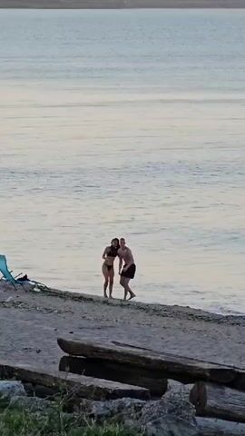Two people, a man and a woman, are standing on a sandy beach near the water's edge. The woman is wearing a dark bikini, and the man is wearing dark shorts. They appear to be interacting, possibly talking or dancing, with the waves gently lapping at their feet. To the left of the couple, a blue beach chair is visible. The beach is littered with driftwood logs, and some green foliage is present in the foreground. The scene is set against a backdrop of calm, rippling water that stretches to the horizon under a pale, overcast sky, suggesting it might be early morning or late evening. The overall mood seems peaceful and relaxed. The location is identified as Shabla, Bulgaria. There is no visible text in the media.