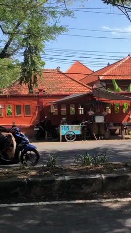 The video opens with a close-up shot of a blue motorcycle parked on the left side of the frame. The motorcycle is partially obscured by a dark object in the foreground. In the background, a red building with orange tiled roofs is visible. The building has several windows and appears to be a shop or restaurant. A small blue cart with a white sign is positioned in front of the building. Two people are sitting behind the cart, and a third person is standing near it. In front of the building, a black motorcycle is parked, and a person is sitting on it. The street is paved, and a sidewalk with a low concrete barrier separates it from the building. Trees and power lines are visible in the background. The scene appears to be a street in a warm, sunny climate.