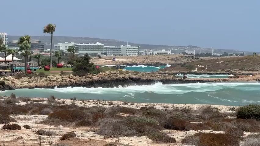 The video shows a coastal landscape with rough seas and a rocky shoreline. In the foreground, there are dry, scrubby bushes and light-colored rocks. The middle ground features the turquoise sea with large, white-capped waves crashing against dark, rocky outcrops. In the background, a line of white buildings, likely hotels or resorts, stretches along the coast under a hazy sky. Palm trees are visible among the buildings, and some people can be seen on the rocky areas near the water. The overall impression is a windy day at a Mediterranean coastal resort.