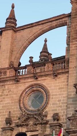 The video shows a close-up view of the exterior of a stone building, likely a cathedral or church, with ornate architectural details. The facade is made of large, rectangular stone blocks. In the center of the frame is a round stained-glass window with intricate tracery. Above the window, there is a balcony with a decorative stone railing. Archways and spires are visible at the top of the structure. The sky in the background is a pale blue. The lighting suggests it is either early morning or late afternoon, with a warm glow on the stone. At the very bottom of the frame, the tops of the heads of two individuals are visible, and a small portion of what appears to be a flag with red, yellow, and green stripes can be seen. The overall impression is of a historic and grand architectural edifice.