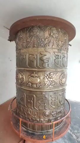 A stationary shot captures a large, ornate Tibetan prayer wheel. The wheel is cylindrical and made of a metallic material, likely bronze, with intricate carvings and inscriptions. The top and bottom rims of the wheel appear to be wooden, with the top rim being a darker reddish-brown and the bottom rim a bright red metal railing around its base. The wheel itself is adorned with multiple horizontal bands of relief carvings. The uppermost band shows decorative floral motifs and figures. Below this are bands of Tibetan script, interspersed with other symbolic imagery, such as flowers and elephants. The prayer wheel is situated indoors, in a room with plain white walls and a light-colored ceiling. A small bell hangs from a wire on the ceiling to the left of the prayer wheel. To the right of the prayer wheel, a large window or glass door reveals an outdoor scene where several people are visible, some appearing to be tourists. The floor beneath the prayer wheel is covered with a red carpet or mat, and the base of the wheel is surrounded by the red metal railing. The lighting in the room is ambient and somewhat dim, with a brighter light source outside the window. The camera is positioned at eye level, looking slightly upwards at the prayer wheel. The video is a single, static shot with no movement of the camera or the subject.