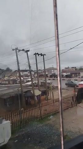 The video shows a street scene with overcast skies. In the foreground, there is a wooden fence and some gravel. Beyond the fence, there are several ramshackle structures, some with corrugated metal roofs and what appear to be tarpaulins. Electrical poles with wires crisscross the scene. In the background, there is a road with several vehicles, including cars and what might be a bus or van. There are also some buildings and more informal market stalls. A few people are visible walking on the street. The overall impression is of a developing urban or peri-urban environment.