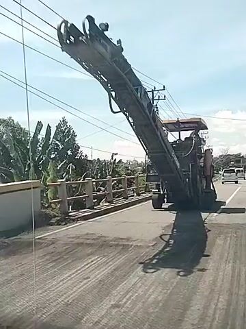 A video shows an enormous asphalt-cutting milling machine crossing a bridge. The machine is tan and black. It is very tall and has a long conveyor belt that stretches high over the bridge. The vehicle appears to be traveling on a paved road lined with trees, utility poles, and wires. The sky is a hazy blue. The video is likely taken from inside a moving car.