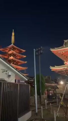 Here's a description of the video:

The video shows an evening view of the Senso-ji Temple in Tokyo, Japan. The camera pans across the temple grounds, showcasing the five-story pagoda and the large Kaminarimon gate. The gate is illuminated and decorated with large paper lanterns. There are many people walking around, appearing to be tourists or visitors. The sky is dark blue.