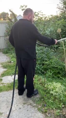 A man wearing a black shirt and black pants is watering plants in a garden. He is holding a black hose with a spray nozzle, and water is spraying onto pink and purple flowers. The garden is filled with green bushes and some plants with light green leaves. The ground is covered with gravel and grass. The sky is visible in the background and appears to be overcast.
