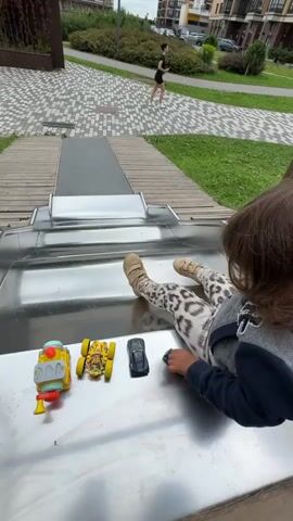 A child, seen from the back, sits on a metal slide with toys in front of them. The child is wearing a gray hooded sweatshirt, leopard-print leggings, and tan socks. The toys include a yellow train, a yellow toy car with a driver, a black toy car, and a black toy with a silver stripe. The slide is part of a playground with a tiled walkway and grassy areas visible in the background. Two apartment buildings are also visible in the distance, along with a person walking on the path. The timecode indicates the video is very short, likely a few seconds.