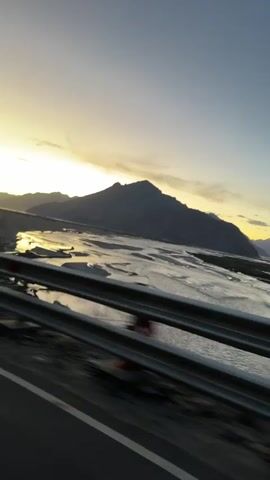 A video shows a person driving along a highway. The landscape outside the car window consists of a large riverbed, mountains, and a clear sky. The sun is setting, casting a warm glow over the scene. The driver appears to be talking to someone in the car, as evidenced by the dialogue snippets. The video captures the beauty of the natural landscape and the experience of a road trip.
