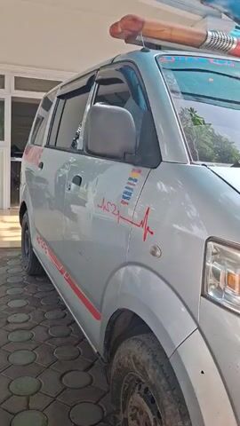 A silver ambulance is parked outdoors on a paved area. The ambulance has the word "AMBULANCE" written on the side, along with a red heart with a heart rate line running through it. The tires on the ambulance are dirty. The background shows a white building with windows and doors. The video is taken during the day.