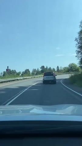 The video shows a view from the interior of a car looking forward, presumably from the dashboard area. The car is traveling on a paved road with two lanes in the visible direction. The road curves to the right. On the left side of the road, there is a guardrail and then a grassy embankment with trees and foliage beyond. On the right side of the road, there is also a grassy area with trees. The sky is clear and blue with a few white clouds.

In the first three seconds, a silver SUV is visible in front of the car, traveling in the same direction and in the right lane. Around the 2-second mark, a large orange and red dump truck appears, passing the SUV and moving into the left lane, overtaking both vehicles. The dump truck is traveling in the opposite direction of the car from which the video is recorded.

After the dump truck passes, the silver SUV continues in the right lane, following the curve of the road. The video appears to be shot during daylight with sunny conditions, as evidenced by the clear sky and shadows.