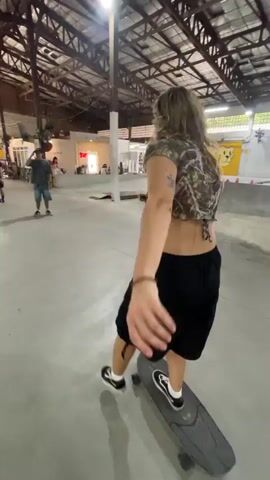 In a skateboard park under a metal-framed roof, a female with long blonde hair wearing a cropped top and black skirt skateboards towards a small ramp. She passes another skateboarder wearing a white t-shirt and jeans. The park contains ramps and other structures. There are a few people standing around in the background. Banners and posters are visible on the walls.