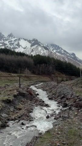 The video shows a rocky stream flowing downstream against a backdrop of snow-capped mountains under a cloudy sky. The stream bed is filled with rocks of various sizes and colors. The water is clear and flowing rapidly. On either side of the stream, there is a mix of grass and rocky terrain. A line of bare trees leads to a dense evergreen forest. A weathered, brown metal pipe crosses the stream. The mountains in the background are partially obscured by clouds.