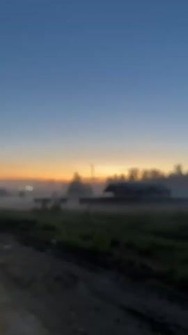 The video shows a rural landscape at dawn. The sky transitions from a deep blue at the top to an orange glow on the horizon. In the foreground, there is a dirt road with visible tire tracks. Beyond the road, there is a field of green grass. A building is visible in the distance, partially obscured by fog. Trees surround the building, further contributing to the hazy atmosphere. A lamppost stands tall, its light barely visible against the brightening sky. The overall scene is tranquil, evoking a sense of calm and rural beauty.