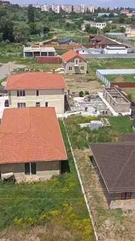 This video appears to be a drone recording of a rural residential development. The scene consists of several houses under construction or recently completed, with red-tiled roofs being a prominent feature. The houses vary in size and style, with some appearing to be single-story bungalows and others multi-story homes. There are also construction materials visible, such as piles of bricks and lumber. The landscape is characterized by rolling hills covered in greenery, with some trees and a body of water in the background. Several dirt roads wind through the development, and utility poles are visible. In the distance, there are some larger, more industrial-looking buildings with arched roofs, possibly greenhouses or storage facilities. A few cars are parked on the property.

The time of day appears to be daytime, with clear, bright lighting, suggesting fair weather. There are no people clearly visible in the footage. The overall impression is one of a developing rural community, possibly in a temperate climate. The drone is moving and panning, providing an aerial perspective of the area. The footage is shot from a relatively high altitude, giving a broad overview of the landscape and the scattered structures.

There is no visible text in the video.