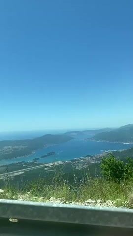 The video pans across a vast, clear blue sky above a coastal landscape. Below the sky is a body of water, likely the sea or a large bay, dotted with islands and peninsulas. Along the coastline, there are indications of human settlements and infrastructure, including buildings, roads, and possibly an airport runway. The foreground shows a metal guardrail and some sparse vegetation, suggesting the view is from a high vantage point, possibly a road winding up a mountainside. The overall impression is of a bright, sunny day with expansive natural beauty and some signs of civilization.