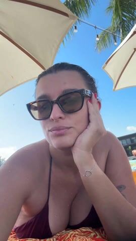 Here is a detailed description of the video:

The video shows a woman in sunglasses relaxing under two beach umbrellas, presumably at a beach or resort. She is wearing a dark bikini top and a silver bracelet with a heart charm on her right wrist. Her hair is slicked back, and she rests her head on her hand, looking directly at the camera. The background includes a bright blue sky, a palm tree, and glimpses of a building and other people. The overall impression is one of relaxation and leisure.