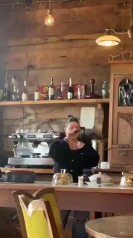 A woman stands behind a cafe counter, facing the camera and gesturing with her hands. She holds a cell phone in her left hand and appears to be listening to or singing along with music playing. The cafe has a rustic decor with wooden walls, shelves, and an old-fashioned cabinet. Shelves behind the counter display bottles and decorations. A coffee machine sits on the counter, along with plates of pastries. Tables and chairs are visible in the foreground.
