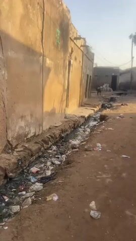 The video shows an outdoor scene in a dusty, underdeveloped area. The foreground is a dirt path with trash scattered across it. To the left, a long, narrow ditch runs alongside a rough, tan-colored building. The ditch is filled with murky water and debris, including plastic bags and other refuse. The building appears to be made of mud or adobe and has several doorways, some of which are open. In the background, more buildings of similar construction are visible, along with a few trees and utility poles with wires. A small, dark-colored animal, possibly a dog, is standing in the middle distance near a pile of white sacks. The lighting suggests it is daytime, with sunlight casting shadows. The overall impression is one of neglect and poor sanitation.