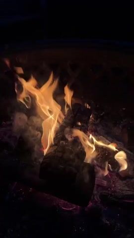 The video displays a close-up view of a bonfire in a fire pit. The fire is actively burning, with flames of various sizes and colors, ranging from bright yellow to orange. Logs of wood are visible in the center of the fire pit, partially consumed by the flames and reduced to glowing embers. The background is dark, suggesting that the video was filmed at night or in a dimly lit environment. A metal grate or the rim of the fire pit is visible at the top of the frame, indicating the confines of the fire pit. The overall impression is one of warmth and flickering light against a dark backdrop.