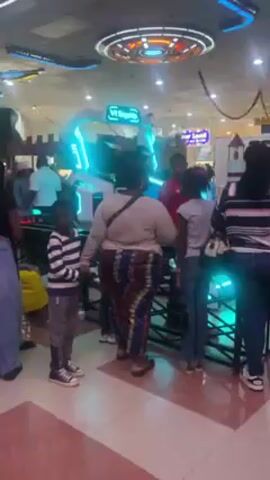 The video shows a group of people standing near a "VR Skyship" virtual reality ride in what appears to be an indoor entertainment center or mall. The VR Skyship is a white structure outlined with bright cyan lights, with people inside wearing VR headsets. The person filming is standing behind a woman with dark hair, a light-colored top, and colorful patterned pants. A young boy in a striped shirt and gray pants is standing to the left of the woman.  Other people, including a young girl with a pink bow in her hair, are standing around the ride. The environment is brightly lit, and there are ceiling decorations visible.