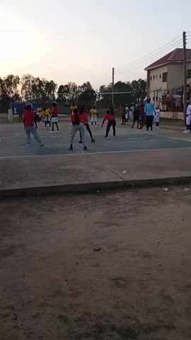 A group of young people in athletic wear are playing volleyball on an outdoor court. The players are mostly wearing red and yellow shirts, with some wearing grey pants. A volleyball is visible in the air, indicating the game is in progress. The court is fenced, and there is a building in the background. The sky is overcast, suggesting it might be late afternoon or early evening. The ground around the court is dirt and gravel.