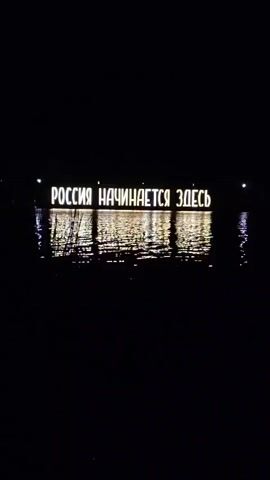 A black and white sign reads "RUSSIA BEGINS HERE" in Russian. The sign is illuminated and reflected in the water below. The scene appears to be outdoors at night.