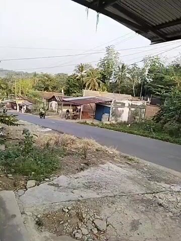 A short video shows a street scene in a rural area, possibly Southeast Asia. The road is paved, with a concrete path on the left. On the right side of the road, there are several small buildings with corrugated metal roofs, some appearing to be shops or stalls. Lush green vegetation and trees are visible in the background, with hills in the distance. The sky is overcast. Several motorcycles are seen riding on the road. One motorcycle is in the foreground on the right, and another is further down the road on the left. There is also a person standing near one of the buildings. The overall impression is of a quiet, somewhat modest, and everyday scene.