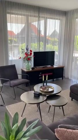 This media file captures a bright, airy living room in Ossenzijl, Netherlands, likely during daylight hours given the natural light streaming through sheer curtains. The scene is set in a modern home, with a focus on a seating area and entertainment setup.

Dominating the visual is a large flat-screen television mounted on a dark wooden media console. The console features a warm-toned wooden top and black cabinet doors, with a shelf displaying a pink book titled "RISK." To the left of the television, a vibrant bouquet of red poppies and blue flowers in a clear green glass vase adds a pop of color and life to the space. A small, dark decorative item, possibly a coaster or a small bowl, sits beside the vase.

In the foreground, a stylish charcoal gray armchair with a metal frame invites relaxation. In front of the armchair and media console, a trio of round coffee tables, varying in size and made of a brass-toned metal, are arranged. The largest table holds a brass tray with two tall green candles in decorative holders and a small clear glass object. A stack of dark coasters rests on the smallest table. The floor is tiled with a neutral gray material, and a light-colored rug is placed beneath the coffee tables.

The background, visible through the sheer white curtains, reveals a glimpse of the exterior. Red-tiled roofs of houses and lush green trees suggest a residential area, possibly near a canal or waterfront, consistent with the Ossenzijl location known for its picturesque waterways. The overall mood is one of calm and contemporary comfort, with the natural light and floral arrangement contributing to a pleasant atmosphere. No people are visible in the frame.