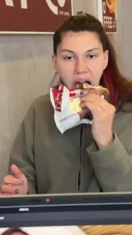 A woman is eating a packaged pastry in what appears to be a coffee shop. She has long brown hair with red highlights and is wearing a gray hoodie. She's sitting at a table with a laptop in front of her, with a portion of the screen visible. The pastry is partially unwrapped, revealing a brown filling. Signs for "COTTI COFFEE" and a graphic for "FFEE" are visible on the wall behind her. She looks directly at the camera while chewing with her eyes looking upward. She seems relaxed and unconcerned while eating. Her left hand has pale, possibly gray, nail polish.