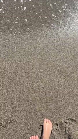 A video shows a pair of feet standing on wet sand as a wave washes over them. The sand is greyish-brown and wet. The waves are white with bubbles and foam. The feet appear to be light-skinned.