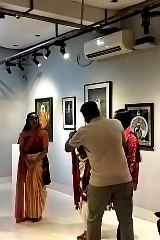 The video shows the inside of an art gallery. A woman in a sari stands in the foreground, facing the camera. A man in a tan shirt adjusts the scarf of a young boy also standing in the foreground. A woman in a red patterned shirt stands behind the man and the boy.  

The walls of the gallery are lined with framed artwork. The lighting is bright, with track lighting affixed to the ceiling. An air conditioning unit is also mounted on the wall. 

Several people are visible in the background, some holding cameras.  The overall impression is of an art exhibition, with someone posing for a photograph.