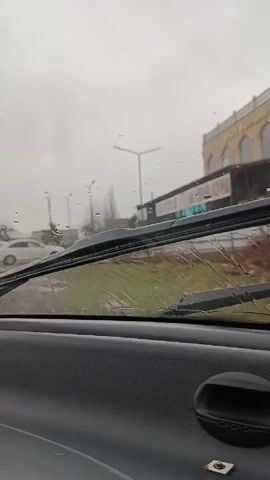 The view from inside a car shows a gray, rainy day through the windshield. The wipers are actively clearing the water. The dashboard is visible, showing a digital clock that reads 12:24. Outside, a street is lined with a grassy median and a building with arched windows. A white car is also parked on the side of the road.