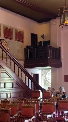 The video shows an interior scene with ornate red chairs arranged in rows. A wooden staircase with a railing is visible on the left, leading up to a balcony with more wooden railings and some electronic equipment. On the right side of the frame, two people are seated in the chairs. A woman in a black top is on the left, with her legs crossed and holding a fan. A man in a patterned shirt is seated next to her on the right. The walls are painted a light color, and there are framed artworks and a decorative element with a circular design visible in the background. The lighting suggests an indoor environment, possibly a hall or a theater.