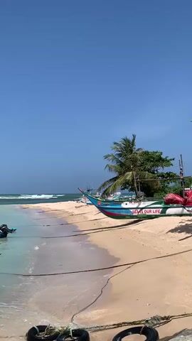 A vertical video shows a tropical beach scene. The foreground features sand and the gentle lapping of waves. Two black tires are partially visible in the bottom left and right corners, connected by ropes. A fishing boat, painted with blue, green, and red accents, is pulled up onto the sand. The boat has the words "SEAS OUR LIFE" written on its side. A palm tree and other greenery are located behind the boat. In the background, the ocean stretches to the horizon under a clear blue sky. A bird is visible flying in the sky. The overall atmosphere is peaceful and serene.