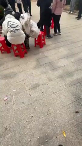 A person is recording a video of themselves and others standing in an outdoor plaza, likely in a residential area in China. The plaza has brickwork on the ground and a few small landscaped areas. There are multiple people in the background, standing or sitting on red plastic stools, potentially waiting for an event. A small, single-story building with a red roof can be seen behind the group of people. The video has a lot of camera movement. There is loud and distracting noise throughout the clip.