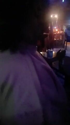 The video shows a dark, blurry scene, appearing to be inside a dimly lit establishment, possibly a bar or lounge. There are tables and chairs in the background, with some people seated and standing. String lights hang above, providing some illumination. In the foreground, a blurry purple object takes up much of the view. A person’s arm can be seen moving around, holding a container, likely a beverage. There's a sense of activity in the background, as people seem to be conversing and moving around. The overall ambiance suggests a nighttime social gathering with low lighting and movement.