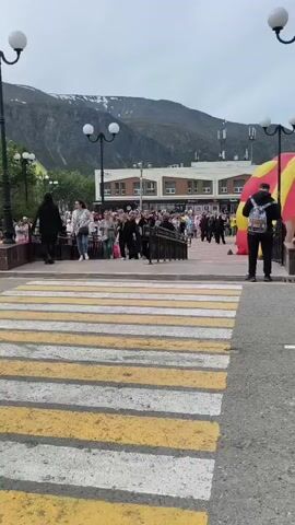 A pedestrian crossing with yellow and white stripes is in the foreground. Beyond the crossing, a crowd of people are gathered. Some people are walking, others are standing. There is a railing with metal posts and a black top. In the background, there is a building with a sign that reads "СУПЕРМАРКЕТ". To the right of the building, there is a large red and yellow inflatable object. The sky is overcast and there are mountains in the distance. A person wearing a black jacket and a backpack is standing on the right side of the pedestrian crossing. Another person wearing a black t-shirt and black pants is walking across the pedestrian crossing.