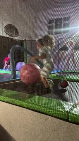 A young girl with blonde hair in a white dress is on a trampoline court with a large pink ball. She is holding a blue curved pole and appears to be swinging it. There is another child in the background wearing a pink shirt and another child in the distance wearing a pink outfit. The environment is a trampoline park with safety netting and a basketball hoop mounted on the wall. The lighting is dim and appears to be artificial.