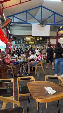 The video pans across a bustling indoor space that appears to be a cafe or restaurant with an attached market area. In the foreground, several wooden tables and chairs are visible, some with black upholstery. On one table, a small plate with a skewer of food and a napkin sits. As the camera moves, people are seen sitting at tables and standing. To the left, a group of people are seated around a table, with one person looking at their phone. Further back, a counter area with shelves stocked with various products is visible, and a fan hangs from the ceiling. In the middle ground, a woman in a black top and blue jeans walks towards the camera, while another woman in a white and black top and denim shorts stands near a shelf. Behind them, more people are seated and standing, with a tall display cooler labeled "SỨA BÒ TƯƠI LONG THÀNH" (Fresh Cow's Milk Long Thanh) and featuring an image of a cow. The environment has a red and blue ceiling structure, with fluorescent lighting. Red and white flags are hung along the upper walls, suggesting a festive or local atmosphere. The floor is a dark, patterned tile. The overall impression is of a lively establishment with both seating and retail elements.