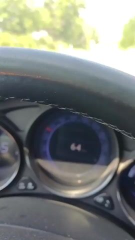 The video shows a close-up of a car's dashboard as it is being driven. The speedometer displays the speed in kilometers per hour, starting at 64 km/h and gradually decreasing to 62 km/h. The tachometer is visible on the left side, and an additional digital display on the right shows the mileage as 1727 and 257125. The steering wheel and parts of the car's interior are also visible. Sunlight streams in from the outside, illuminating the dashboard. The background is blurred, suggesting motion.
