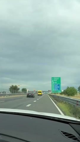 The video captures a first-person view from inside a moving vehicle, traveling along a multi-lane highway under an overcast, cloudy sky. The road features clear white lane markings and a metal guardrail on the right side.

A prominent green highway sign on the right indicates upcoming destinations. The top section of the sign, with an arrow pointing right for an exit, lists:
*   СВИЛЕНГРАД (Svilengrad)
*   БУРГАС (Burgas)

The bottom section of the sign, with an arrow pointing straight for continuing on the highway, lists:
*   ПЛОВДИВ (Plovdiv)
*   ПАМПОРОВО (Pamporovo)
*   СМОЛЯН (Smolyan)

English translations are provided below each Bulgarian name. Below this text, the number "1000" is visible, likely indicating the distance in meters to the exit.

Further down the road, a Shell gas station sign and an overhead McDonald's sign (with its distinctive yellow "M") are visible. Several other vehicles, including a black car and a yellow van, are ahead on the road. The white dashboard of the recording vehicle is visible in the foreground.

The overall setting suggests a journey on a major road near Radinovo, Bulgaria, as indicated by the location context. The time of day appears to be daytime, despite the heavy cloud cover.