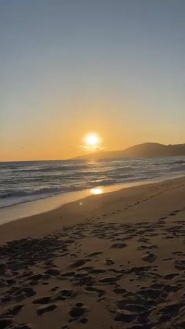 The video shows a beach at sunset. The sun is setting over the ocean, casting a golden glow on the water and sand. The sky is clear and blue with a hint of orange. There are mountains in the distance. Waves are crashing on the shore. There are many footprints in the sand.
